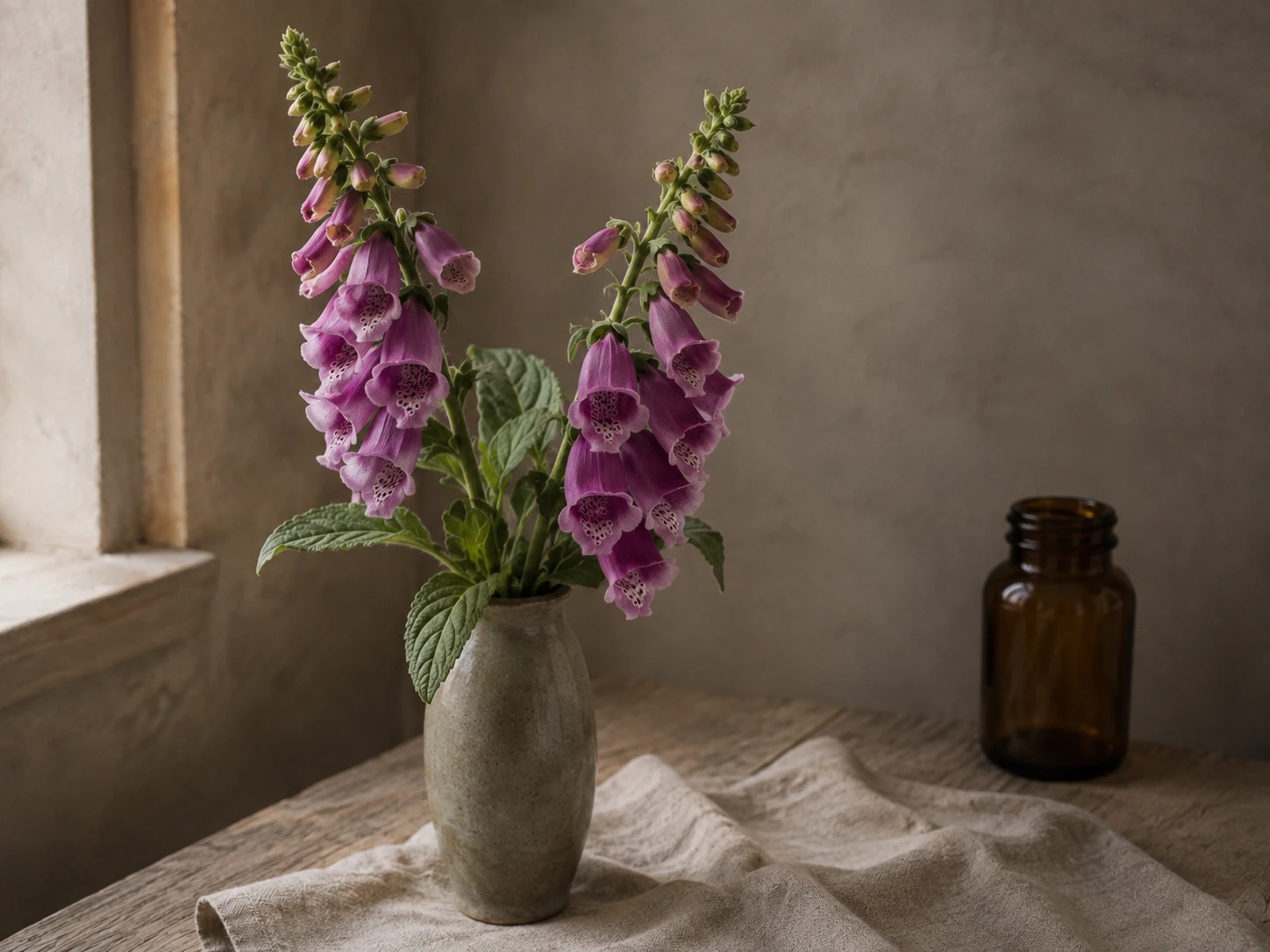 Fingerborgsblommans purpurrosa, fläckiga blomrör och gröna blad i en keramikvas vid ett fönster, med linne och trä.