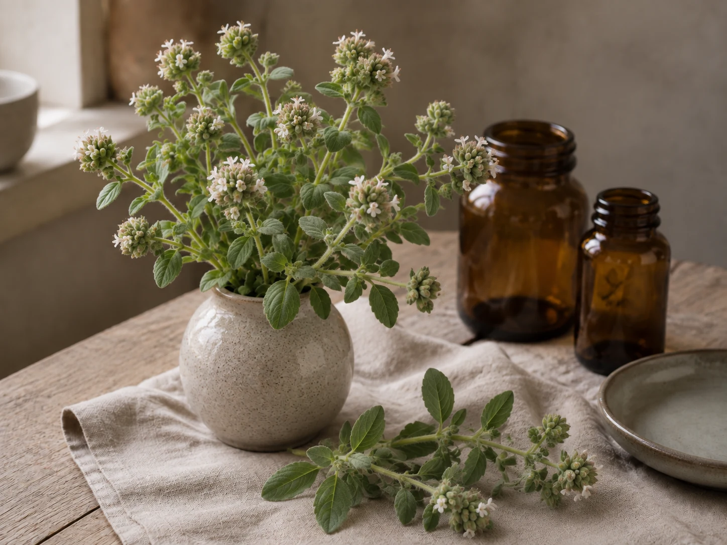 Mejram med små grågröna blad och täta ljusa blomhuvuden i en keramikvas, med en kvist på linneduken, tomma bärnstensfärgade glasburkar och ett tomt keramikfat.